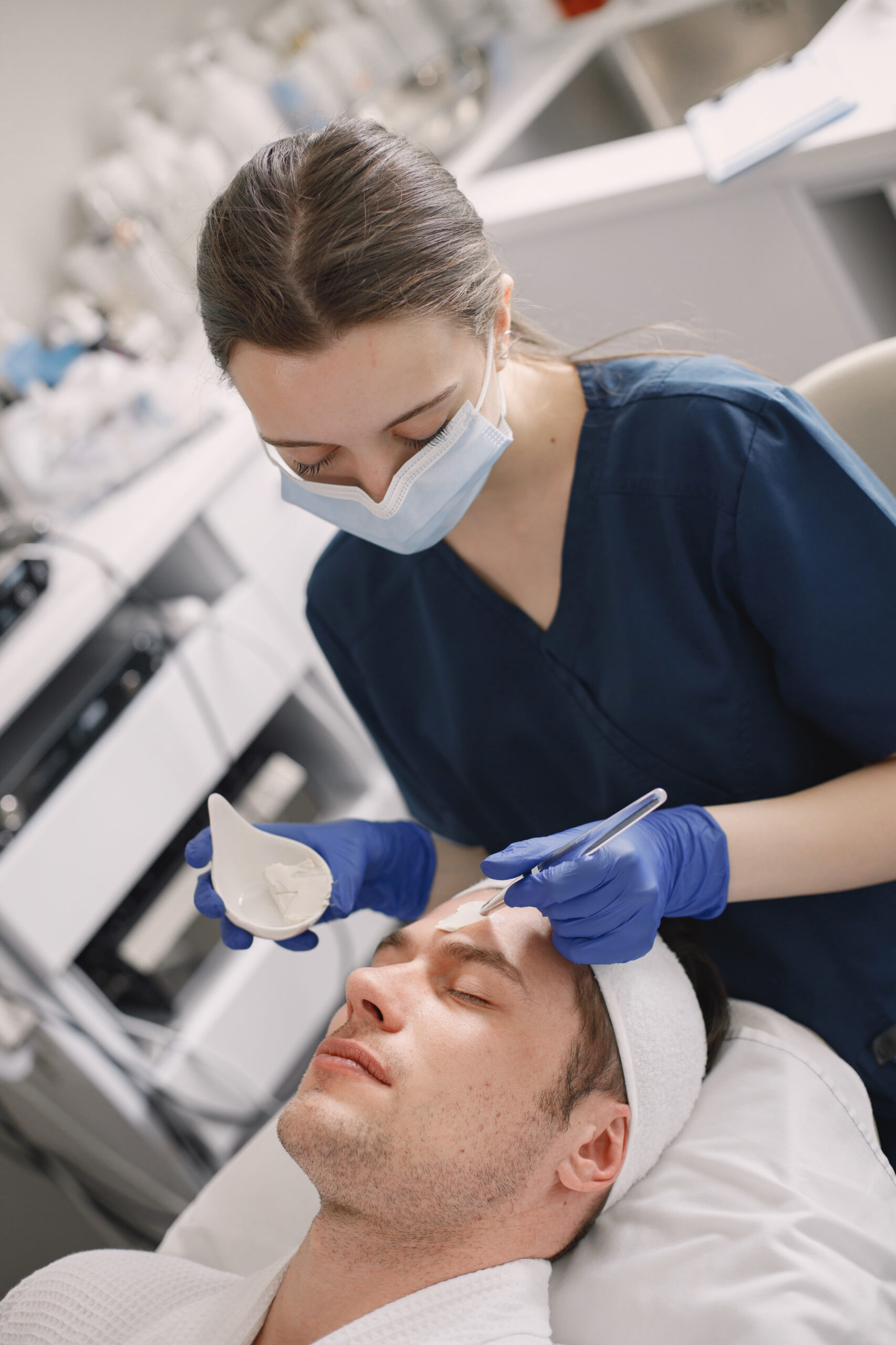 Man laying in cosmetologist cabinet and has a procedure for face skin. Female cosmetologist wearing blue medical costume and face mask. Woman applying a mask on man's face.