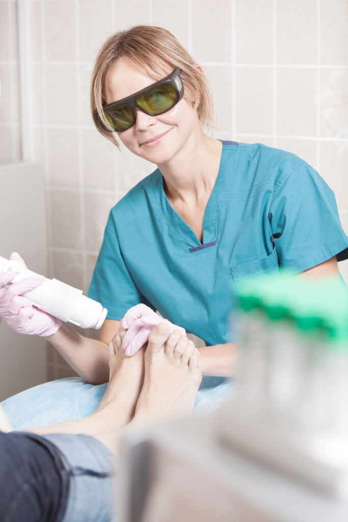 Smiling woman cosmetician in safety glasses working with laser during foot treatment